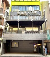 A multi-story building with a green and yellow facade, a covered outdoor seating area on the ground floor, and a visible entrance to a business on the right.
