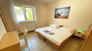Un dormitorio con cama, escritorio, silla, mesita de noche con lámpara y ventana con cortinas.