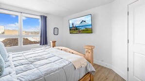 Une chambre à coucher avec un lit, une fenêtre donnant sur un bâtiment et un paysage, un tableau encadré au mur et une porte.