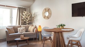 Ein Wohnzimmer mit einer Couch, einem Couchtisch, einem Esstisch mit Stühlen, einer Lampe und einem an der Wand befestigten Fernseher.