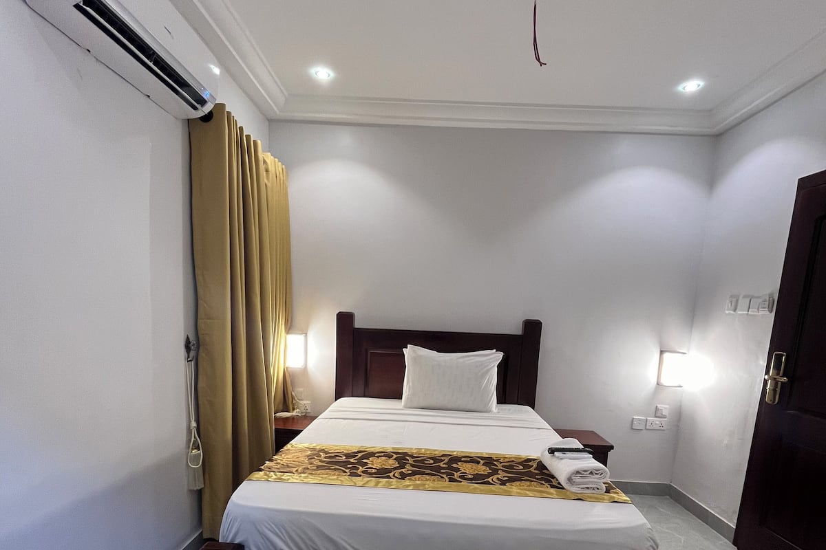 A hotel room with a bed, curtains, a bedside table, a wall-mounted air conditioner, and a door.