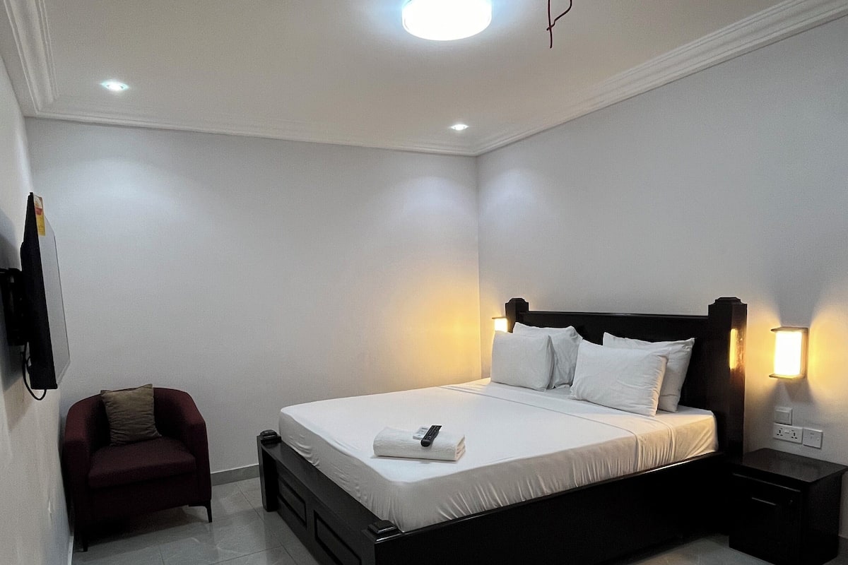 A modern bedroom with a large bed, bedside lamps, a small armchair, and a flat-screen TV mounted on the wall.