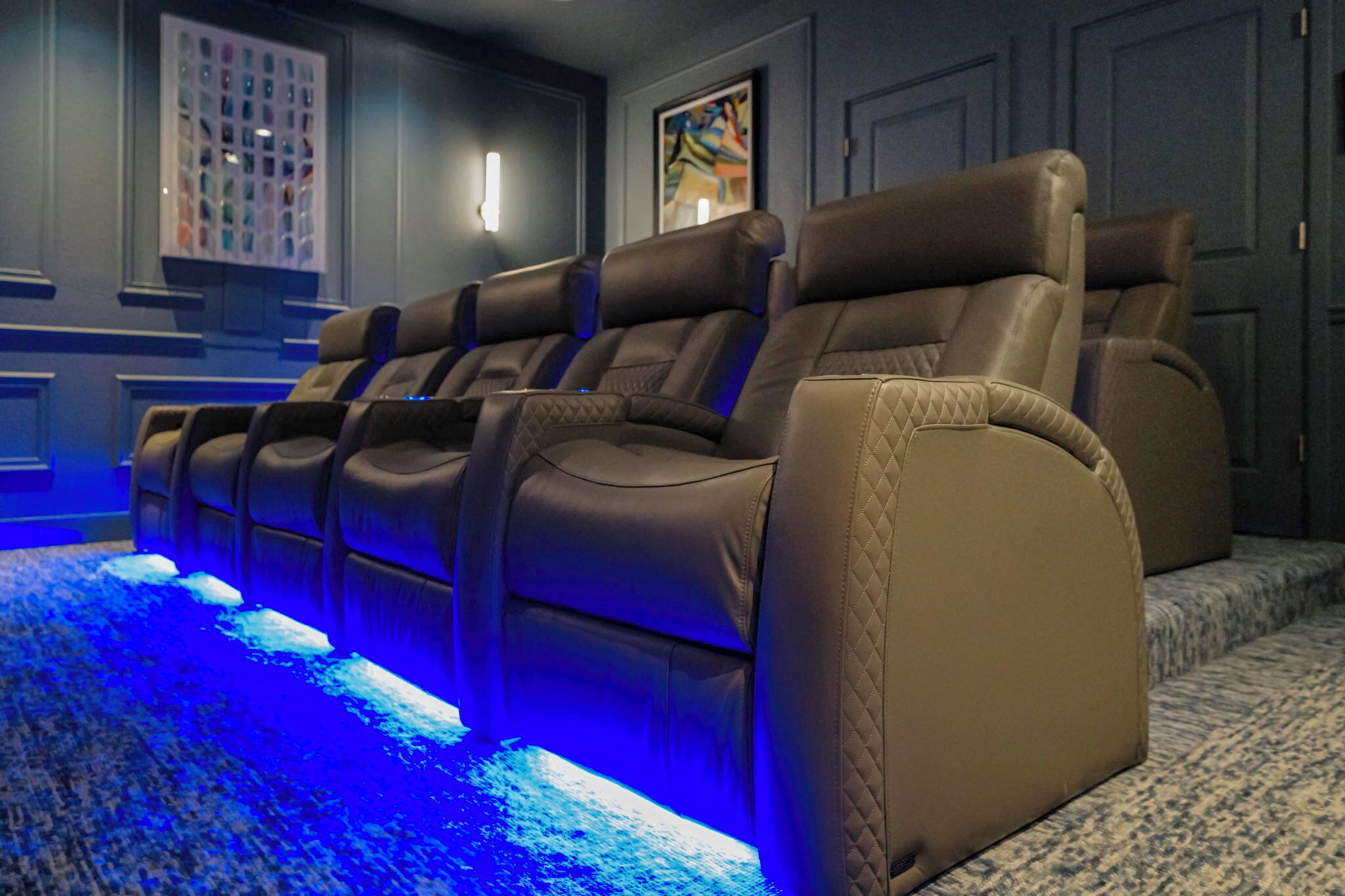 A row of leather recliner chairs with blue ambient lighting, a patterned carpet, and a framed artwork on the wall.