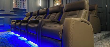 A row of leather recliner chairs with blue ambient lighting, a patterned carpet, and a framed artwork on the wall.