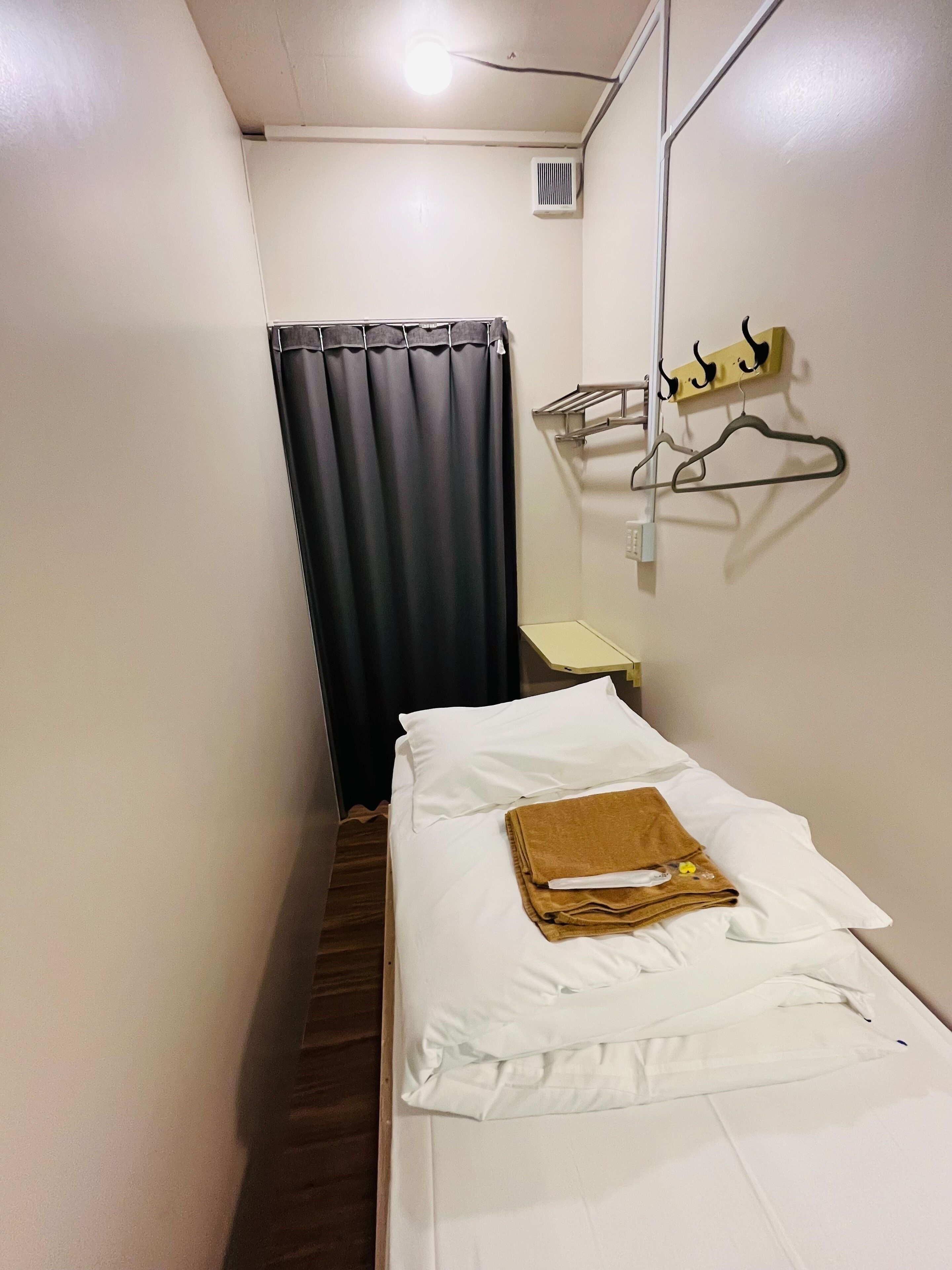 A narrow hotel room with a single bed, a small table, a hanging coat rack, and a curtain.