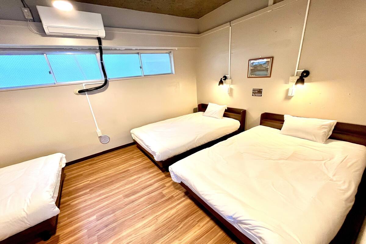A hotel room with two beds, a window, an air conditioning unit, and a wall-mounted light fixture.