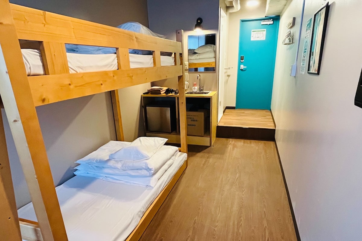 A narrow hallway with bunk beds on one side and a desk with a chair on the other. There is a door with a sign on it and a small step leading to a higher level.