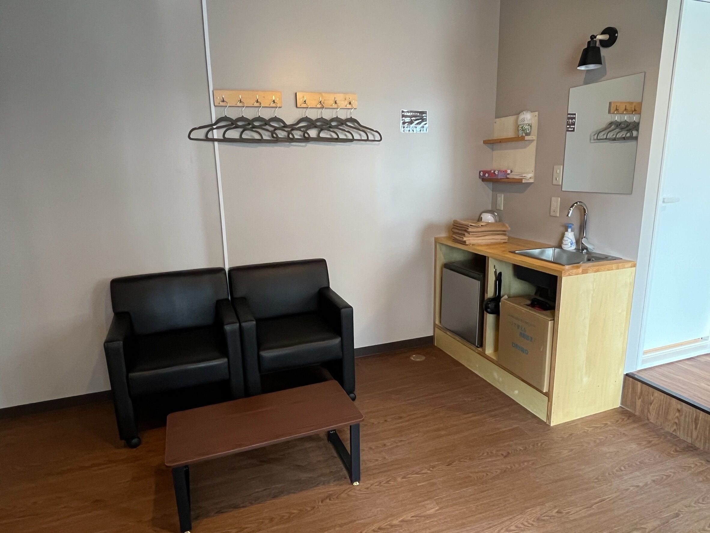 Una habitación moderna con un sofá de cuero negro, un banco de madera, una pequeña kitchenette con un lavamanos y un espejo, y un perchero en la pared.