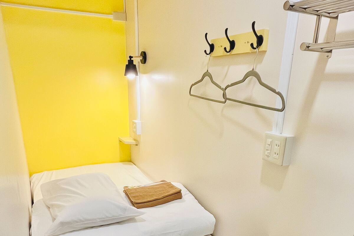 A small, single-bed room with a yellow accent wall, a bedside table, and a wall-mounted hanger.