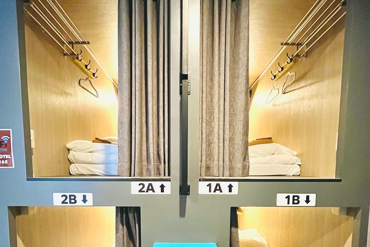 A hotel room with four single beds arranged in a square layout, each with a curtain and a hanging rack. There is a small teal shelf in the center.