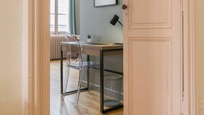 A room with a wooden floor, a desk with a chair, a hanging light fixture, a framed picture on the wall, and a closed door. - Le Bressey  Historic Mansion (Dijon)