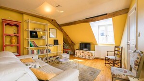 A cozy attic room with a sloped ceiling, a white sofa, a bookshelf, a television, and a window. - Le Panoramique - Exceptional View (Dijon)
