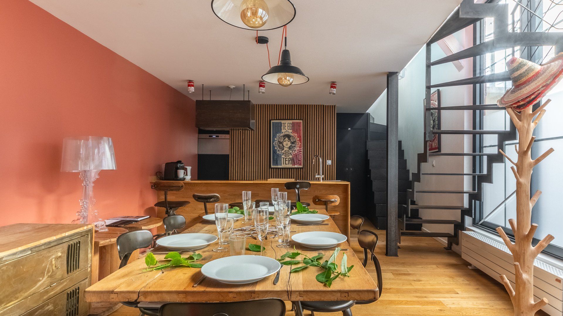 A dining area with a wooden table set for six, a kitchen in the background, and a staircase leading to another level.
