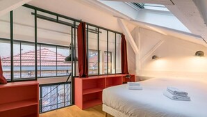 Une chambre moderne avec un puits de lumière, un lit sur lequel sont soigneusement pliées des serviettes, et une étagère rouge.