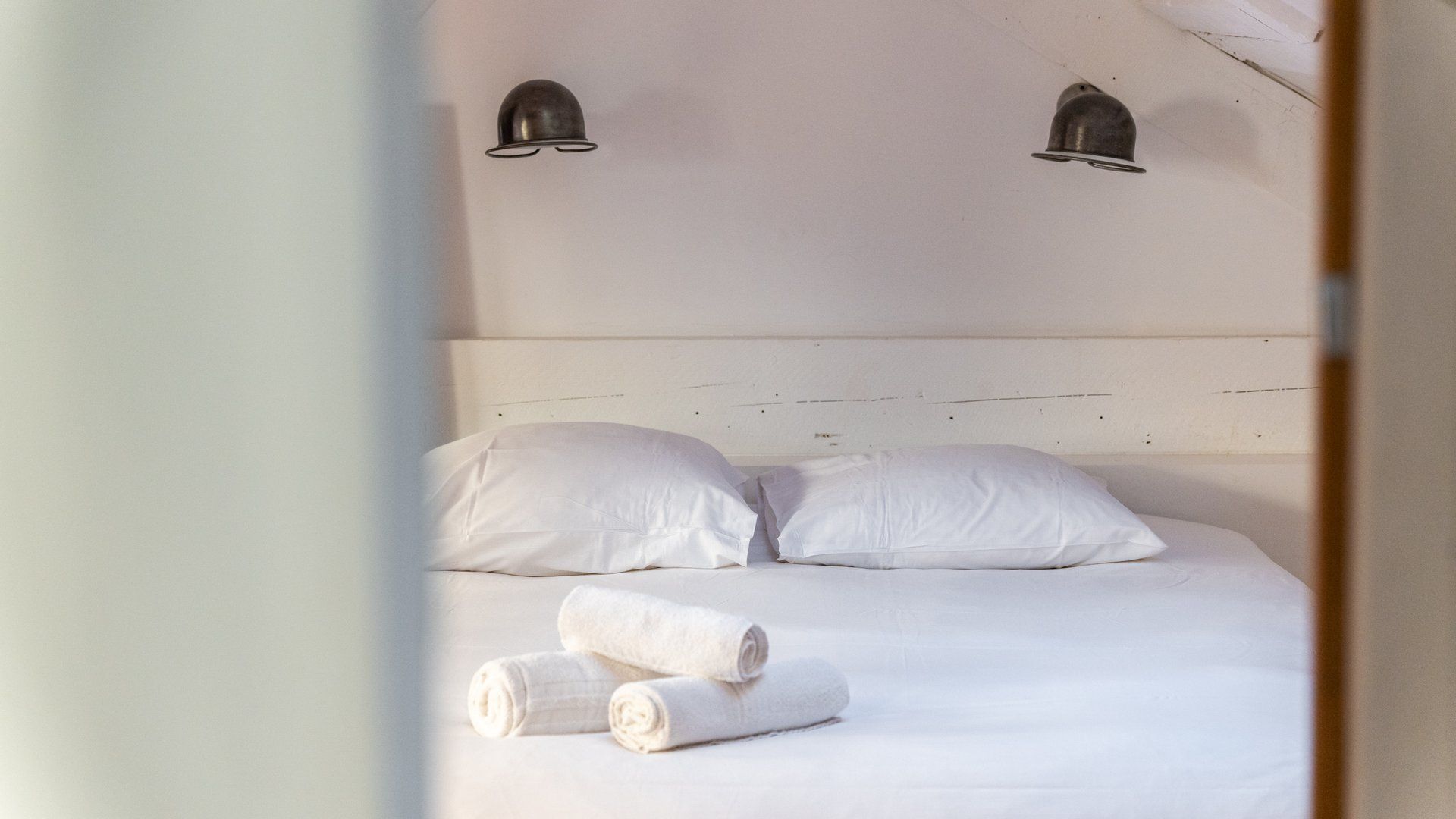 Una cama con ropa de cama blanca y dos toallas enrolladas, un cabecero de madera y dos lámparas fijadas a la pared.