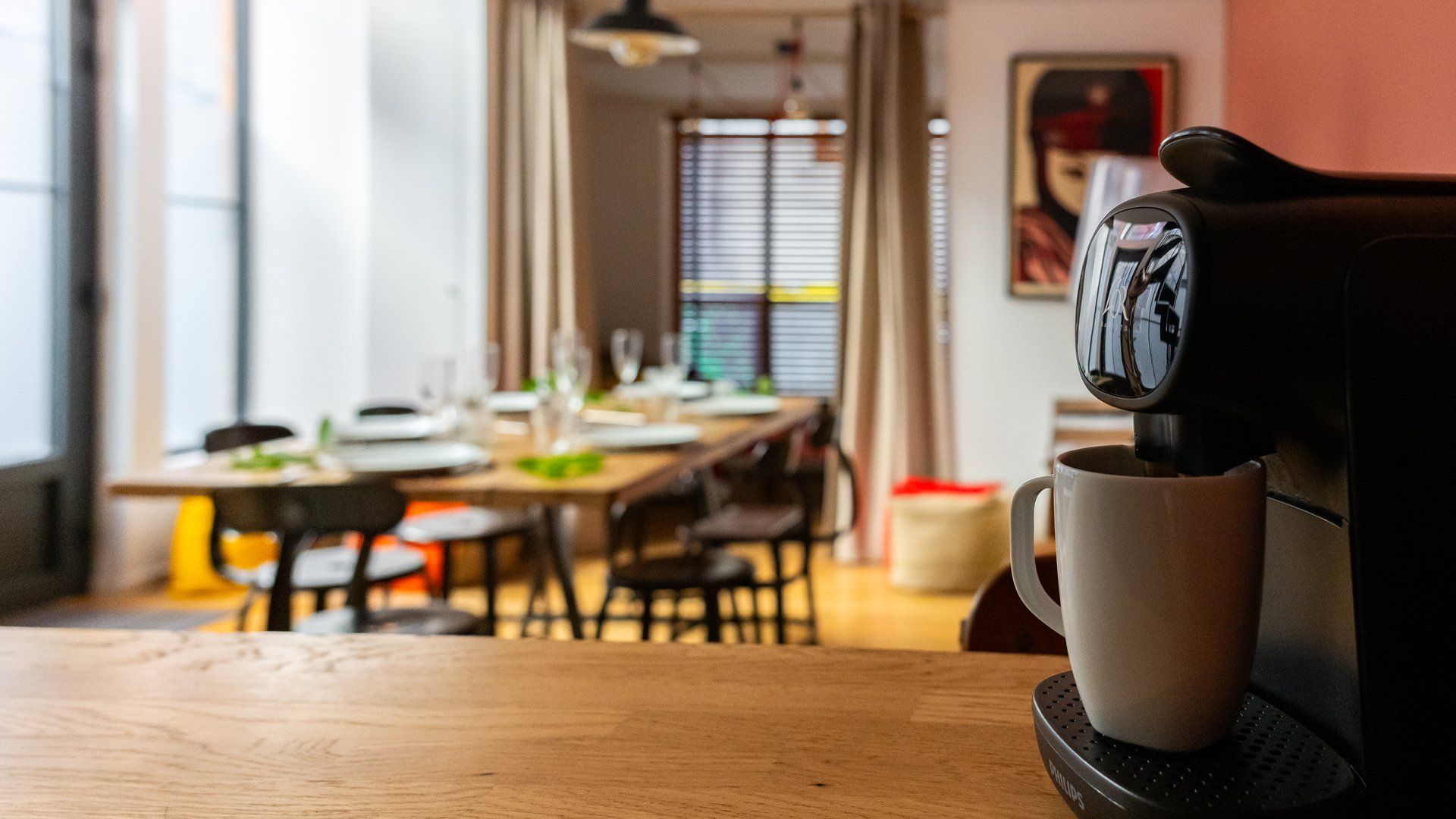 Una cafetera sirviendo café en una taza sobre una mesa, con un comedor al fondo.