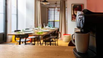 Una cafetera sirviendo café en una taza sobre una mesa, con un comedor al fondo.