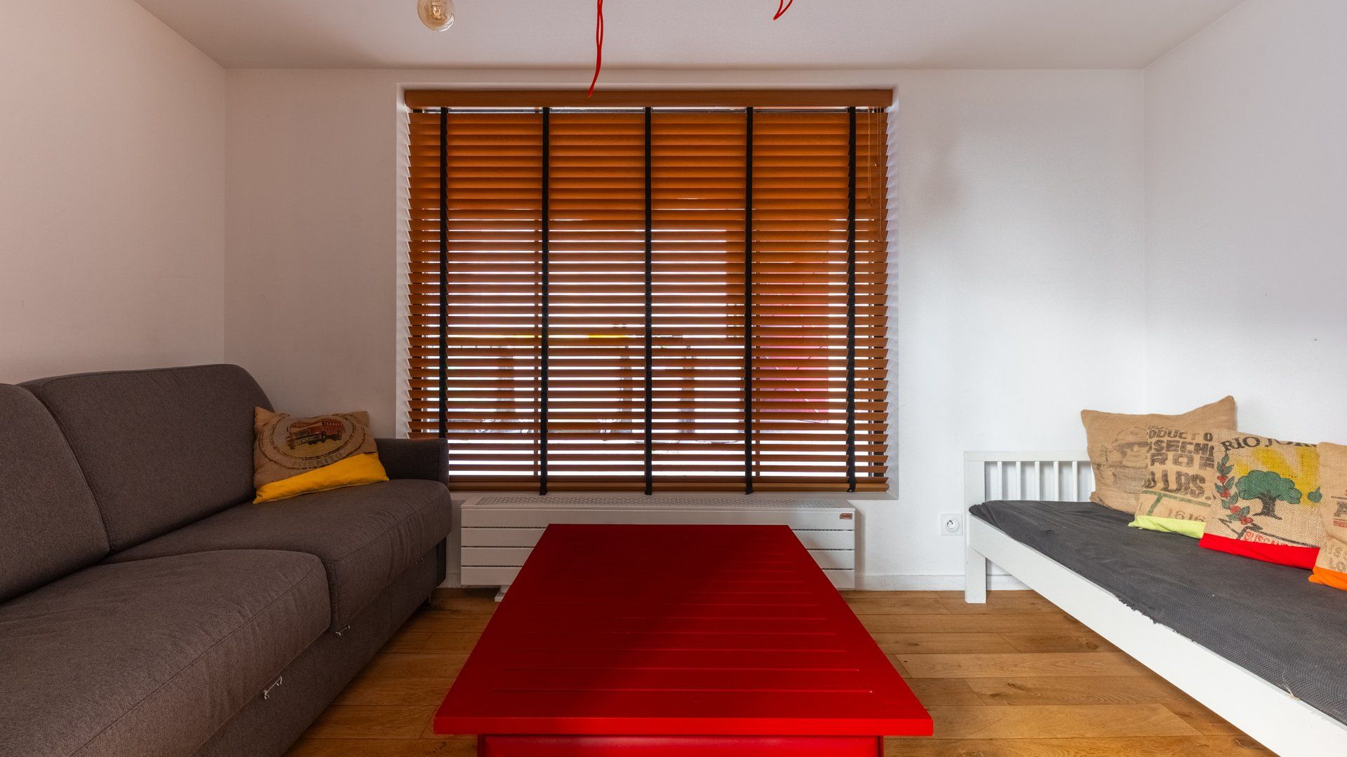En stue med sofa, en rød bænk, et vindue med persienner og en radiator.