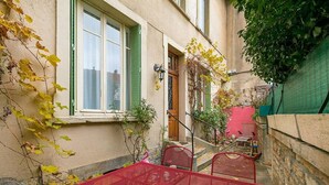 Un patio estrecho con una mesa y sillas de metal rojo, una puerta de madera y una ventana con contraventanas verdes.