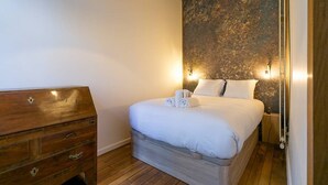 Une chambre avec une commode en bois, un lit recouvert de draps blancs et un mur à texture.