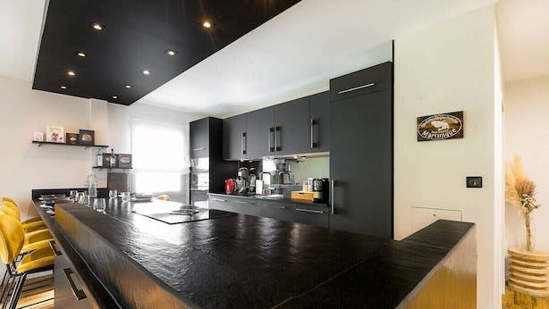 Een moderne keuken met een donker aanrecht, zwarte kastjes en een bar met gele stoelen.