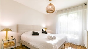 Een hotelkamer met een groot bed, bedlampjes, een nachtkastje en een raam met dunne gordijnen.