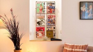 Sala de estar com suporte de revista fixado na parede, planta em vaso, luminária, poltrona com almofada estampada e quadro na parede.