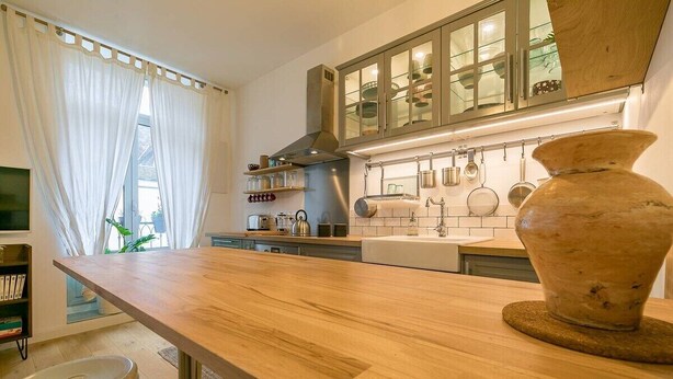 Uma cozinha com bancadas de madeira, um vaso grande e uma janela com cortinas leves.