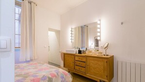 Quarto com cama, toucador de madeira com espelho e iluminação, janela com cortinas e radiador.
