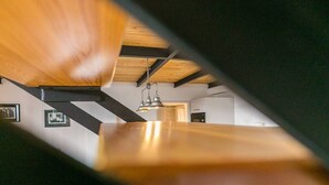 Visão interna de um quarto com vigas de madeira aparente, luminária pendente e quadros nas paredes.