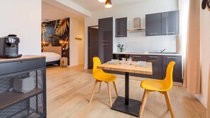 Ett kompakt bostadsutrymme med ett litet kök, en matplats med två gula stolar, en säng och en kaffemaskin.