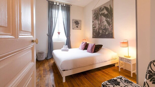 Un dormitorio con cama, lámparas de noche, un radiador, una ventana con cortinas y un cuadro colgado en la pared.