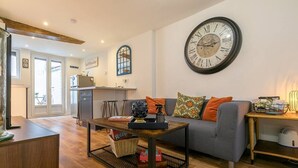 A living room with a sofa, a coffee table, a large wall clock, and a kitchen area in the background. - Château Biek (Dijon)