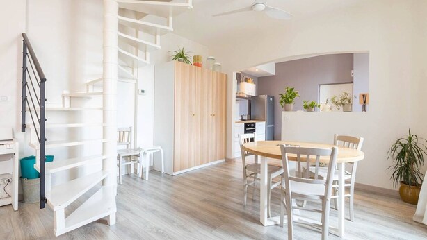 Un interior moderno con una escalera espiral, una zona de comedor con mesa y sillas de madera, y una cocina equipada con electrodomésticos y plantas.