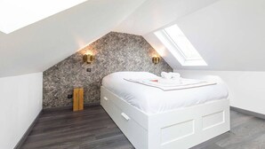 Un dormitorio con techo inclinado, tragaluces, cama con ropa blanca y dos luces fijadas a la pared.