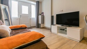 Ett modernt vardagsrum med en platt-TV, en soffa med orange kuddar, en vit TV-bänk och ett fönster med gardiner.