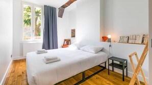 Un dormitorio con cama, una mesita, una silla, un estante con libros y una ventana con cortinas.