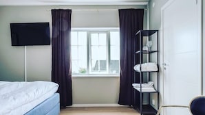 A bedroom with a bed, a window with curtains, a TV mounted on the wall, and a shelf with folded towels. - Hotell MAVI (Viken)