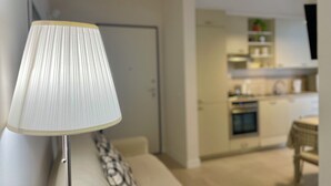 Un salon moderne avec un canapé blanc, un coussin décoratif à motifs et une lampe sur pied. Au fond, on aperçoit une cuisine avec des meubles blancs et un coin repas comprenant une table et des chaises.