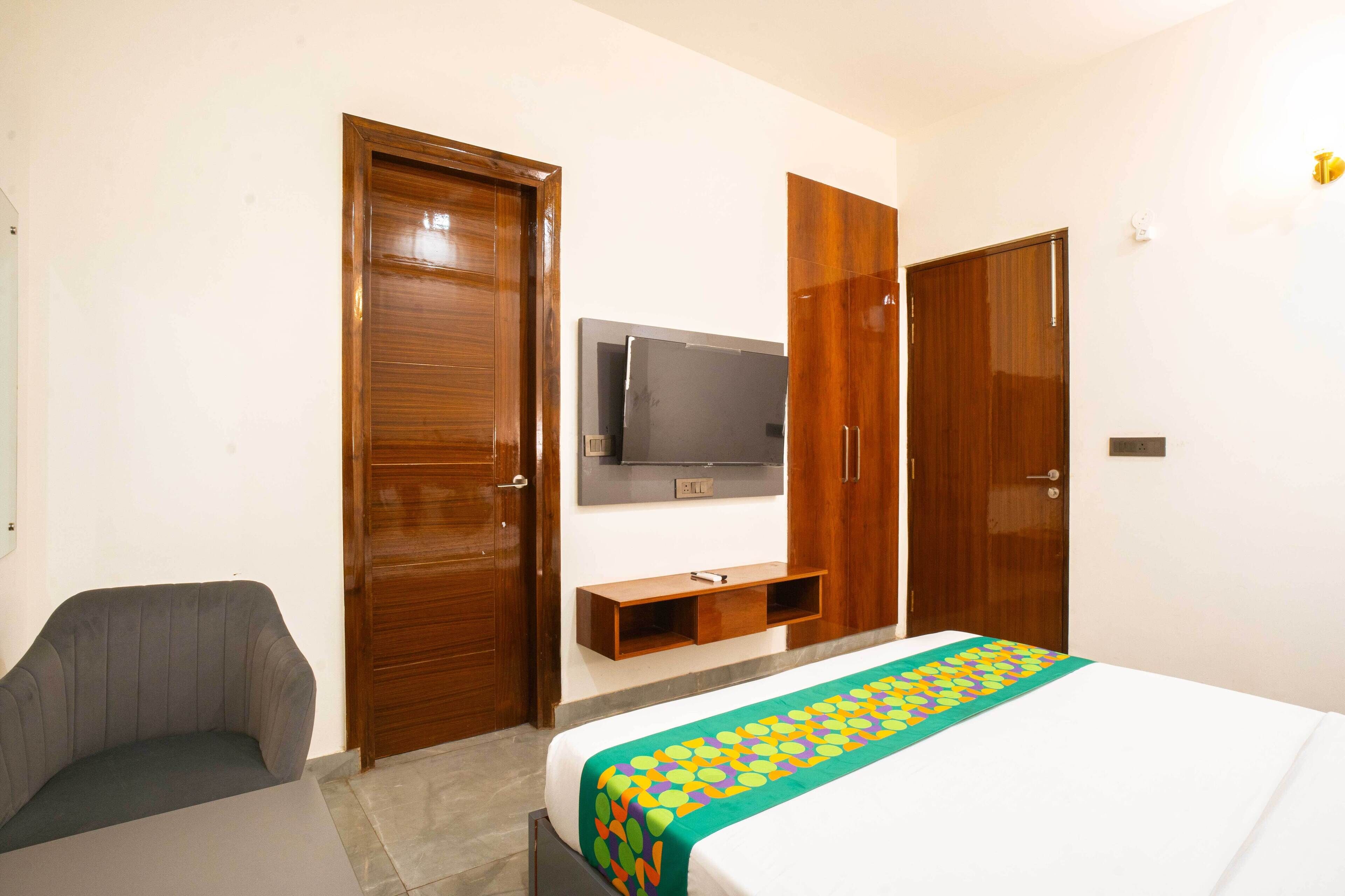 A hotel room with a bed, a TV mounted on the wall, wooden wardrobes, a grey armchair, and a small wooden shelf.