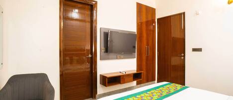 A hotel room with a bed, a TV mounted on the wall, wooden wardrobes, a grey armchair, and a small wooden shelf.