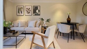 Una sala de estar moderna con sofá, mesa de centro y sillas. Hay obras de arte enmarcadas en la pared y un jarrón con flores sobre una mesita auxiliar.