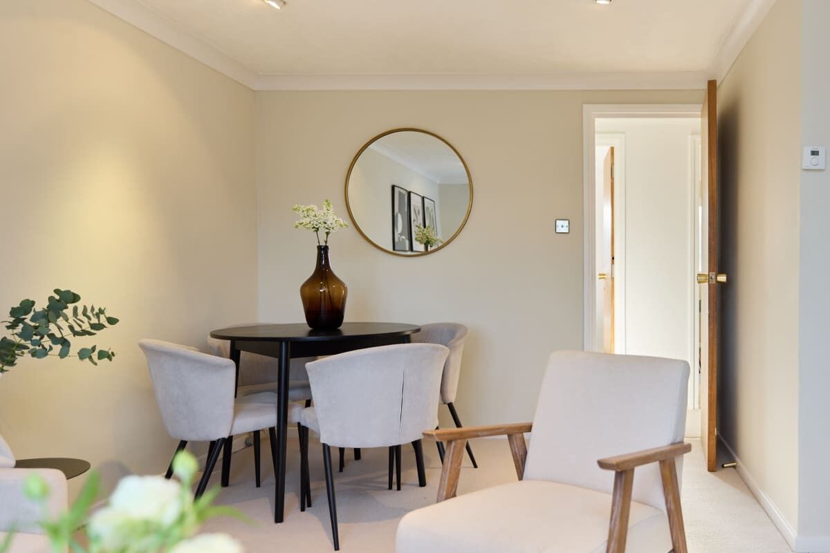 A dining area with a round table, four chairs, a vase with flowers, and a mirror on the wall.