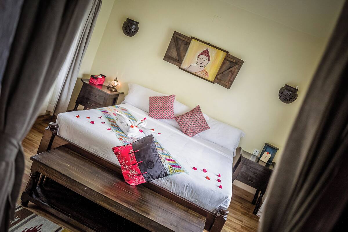 A neatly made bed with patterned pillows, a wooden nightstand with a lamp, and a wall-mounted Buddha painting.