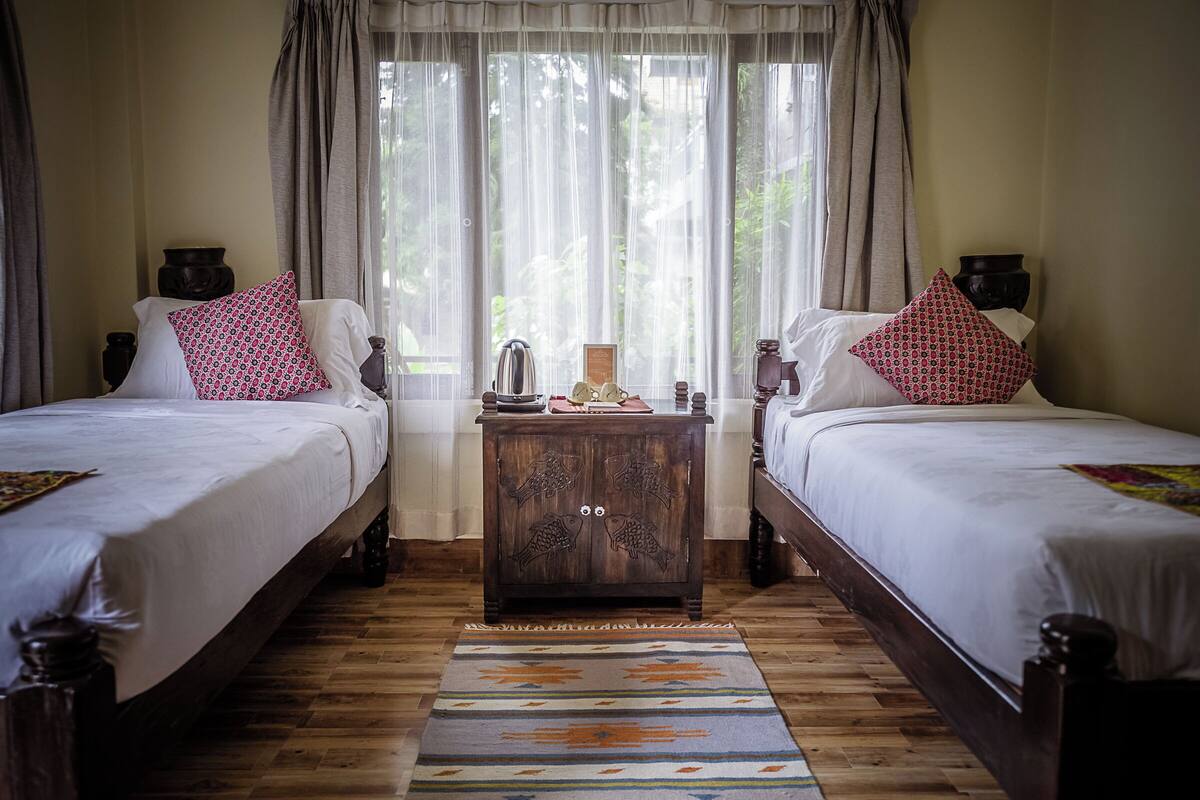 A room with two wooden beds, a nightstand with a lamp, and a window with sheer curtains.