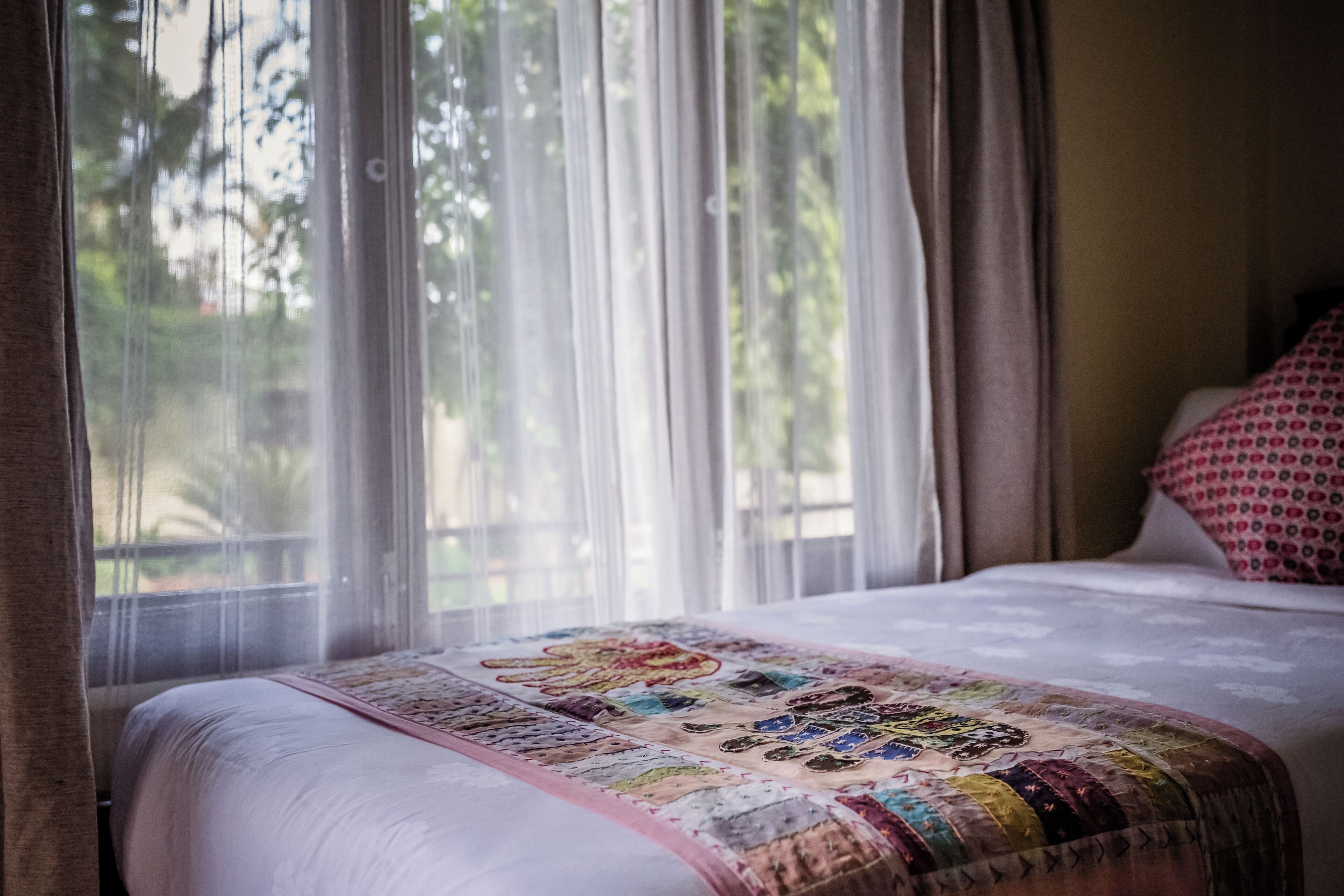 Una cama con colcha, una ventana con cortinas translúcidas y vistas al exterior con vegetación.