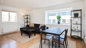 A modern living room with a black sofa, a dining table with chairs, a floor lamp, a bookshelf, and a large window with a view of buildings.