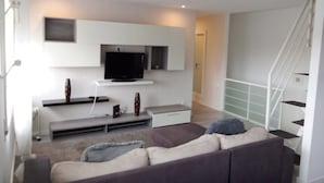A modern living room with a flat-screen TV mounted on a white wall, a grey sofa, a small coffee table, and a staircase to the right. - Miramar Terrace First Line Beach View Villa (Vila Nova de Gaia)