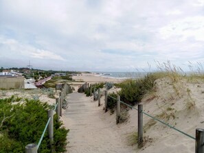 A sandy pathway leading to a beach with a view of the ocean and distant buildings.