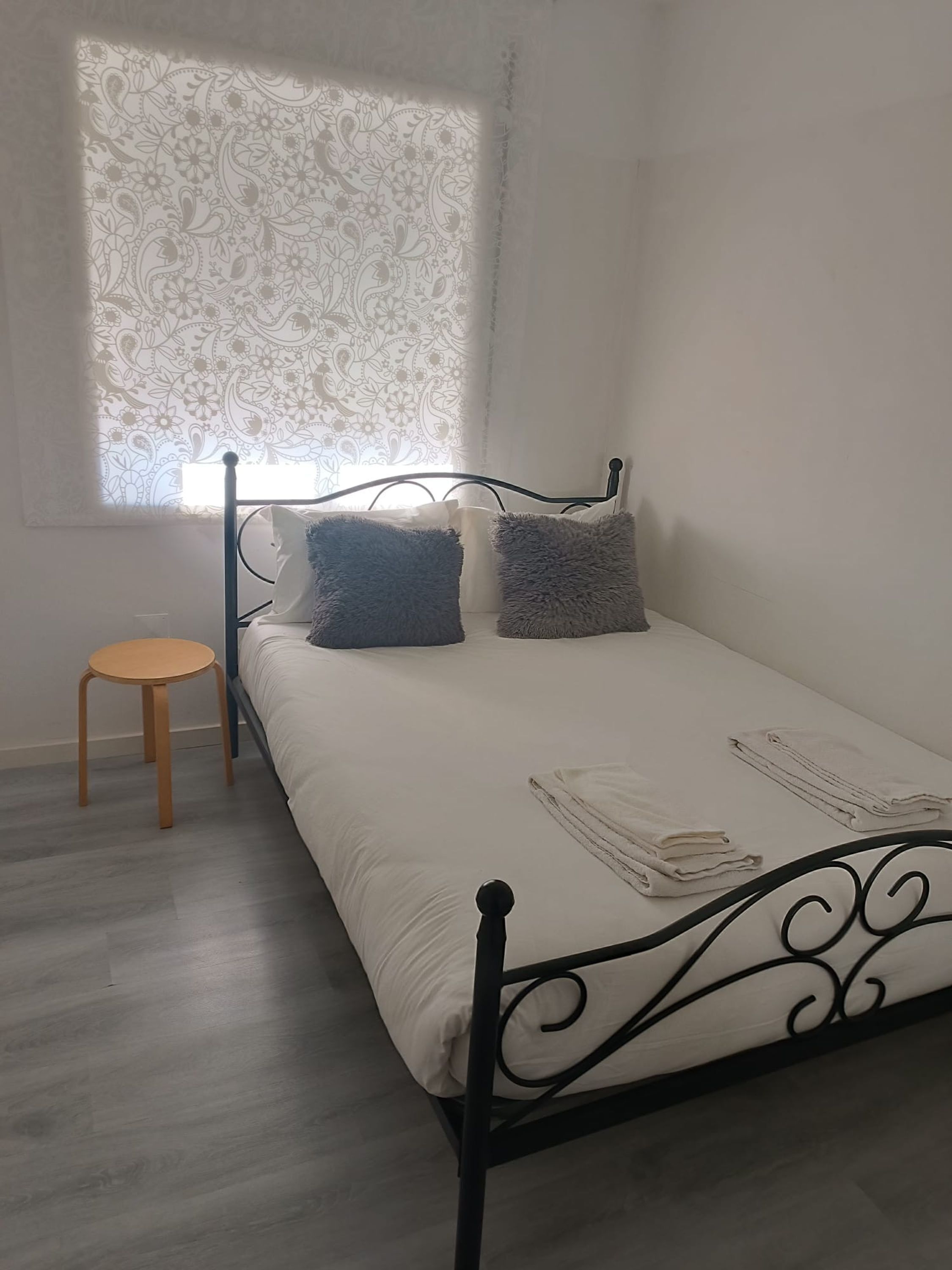 A single bed with a metal frame, a small wooden stool, and a window with a floral pattern.